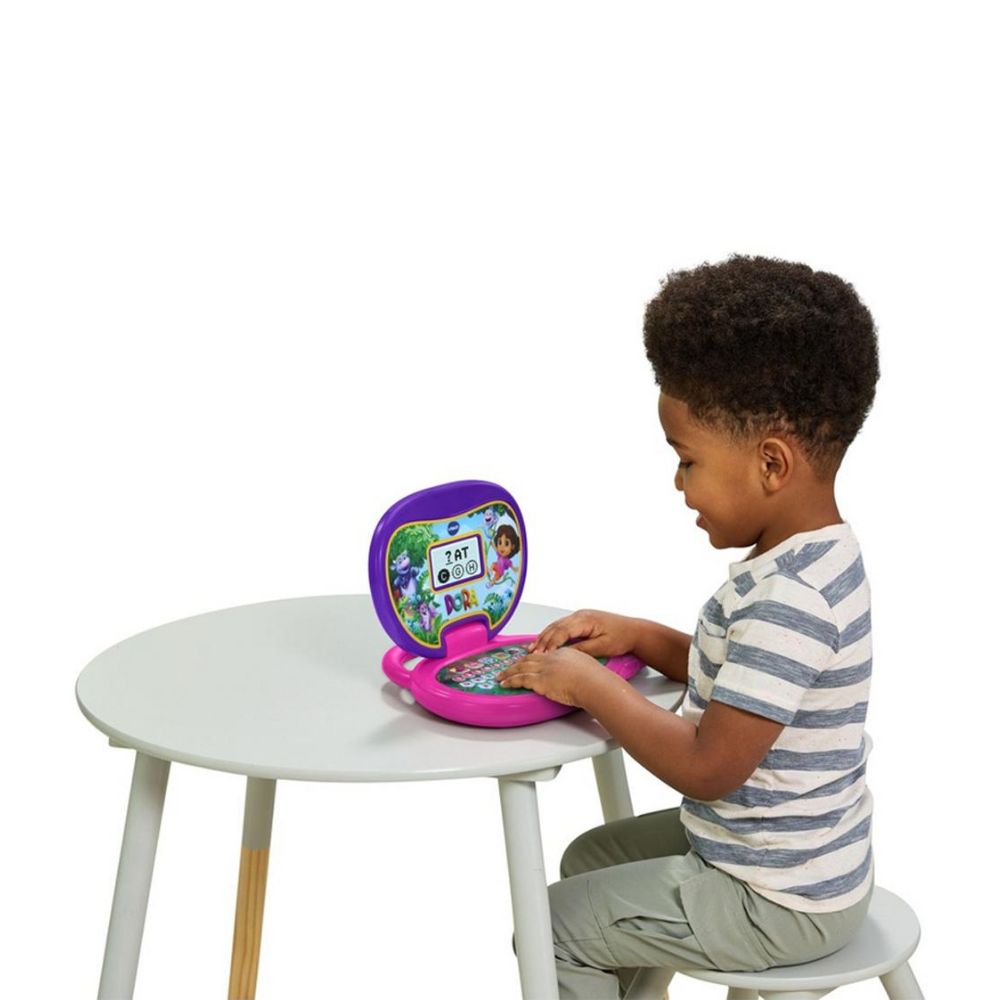 A young child sitting on a small white table, using a colorful pink and purple toy laptop computer against a plain white background.