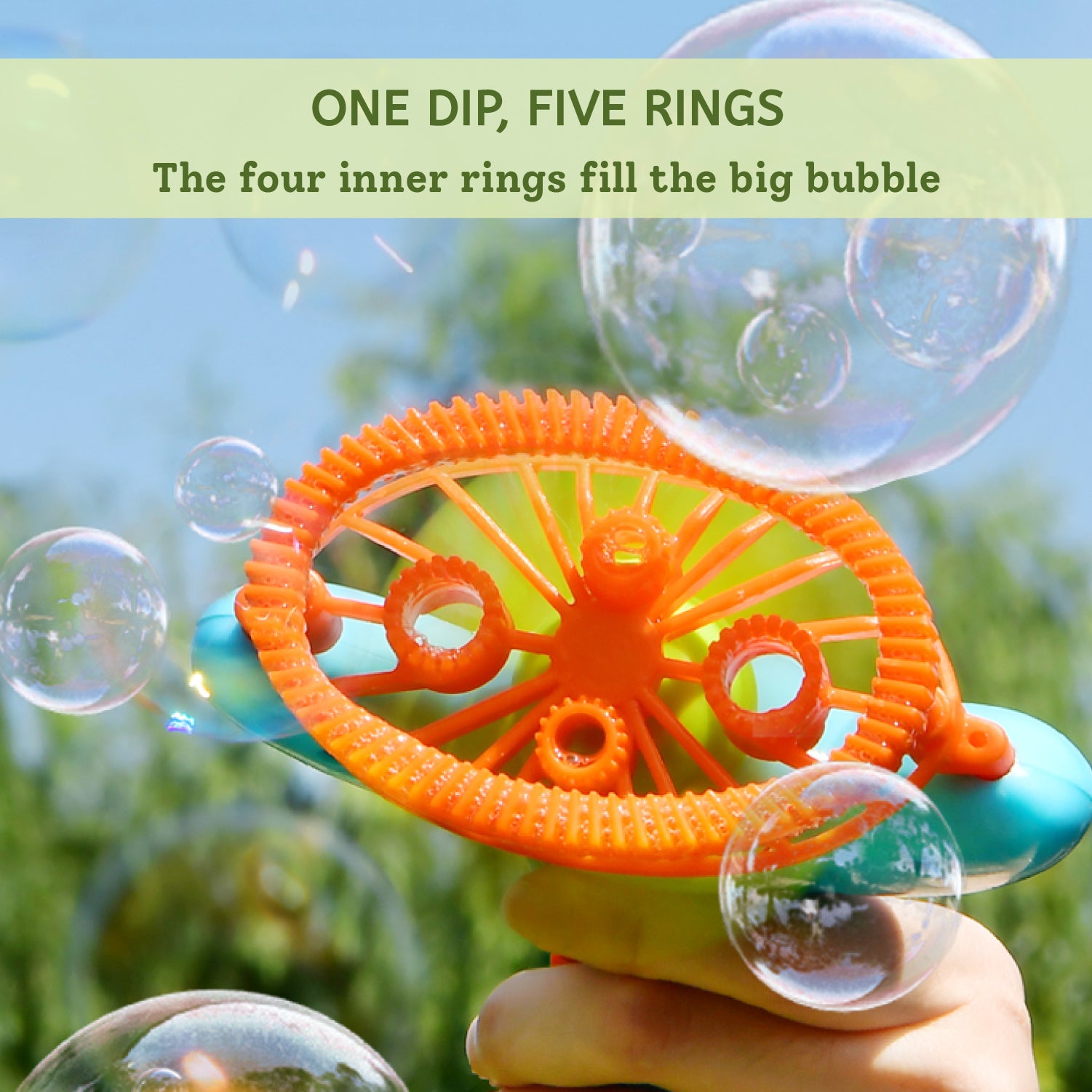 An orange plastic bubble wand with five rings being held by a hand, surrounded by floating bubbles against a blurred green outdoor background. Text at the top reads 'ONE DIP, FIVE RINGS The four inner rings fill the big bubble'.