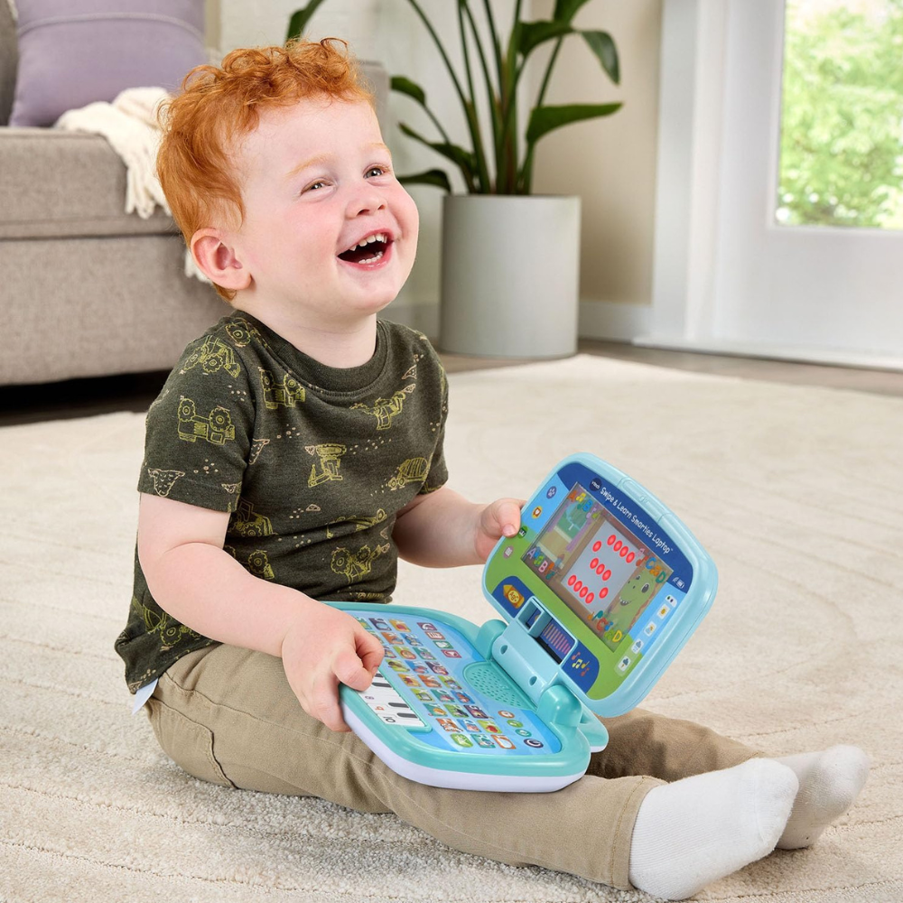 A young toddler with red hair sits on a light beige carpet in a bright living room, smiling and holding a toy laptop learning machine. The toy has a light blue and green design with colorful buttons and a small screen.