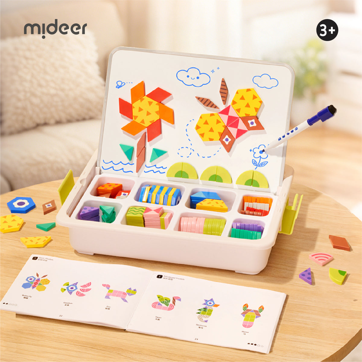 A children's magnetic building and drawing toy set displayed on a wooden table in a bright indoor room. The open white case shows colorful geometric pieces arranged in compartments and a white writing board with drawings of a windmill and star.