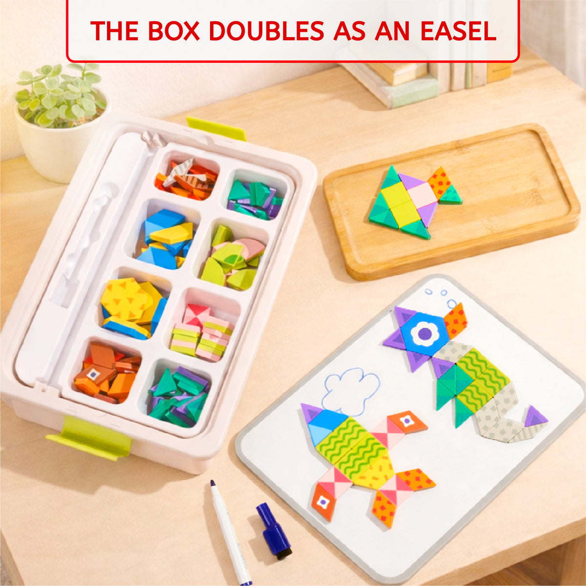 A children's folding paper toy set displayed on a light wood table, including a white storage box with compartments holding colorful folded pieces, a wooden board with a paper fish, a white magnetic-style board with more paper fish, a marker, and a text banner at the top reading 'THE BOX DOUBLES AS AN EASEL'.