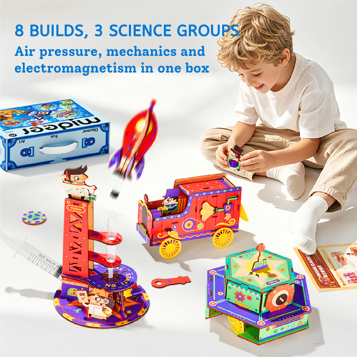 A child sits on the floor playing with a colorful magnetic science toy set on a white studio background. The set includes a blue storage box, a red rocket, a red toy truck, a purple and orange tower structure, and a green and purple box with a face panel.