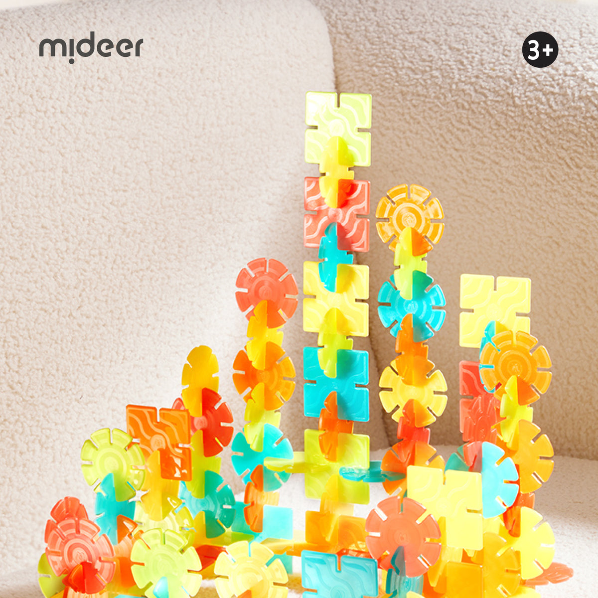 A set of colorful plastic building blocks with interlocking shapes in yellow, orange, teal, and red, arranged on a beige textured fabric surface. The brand name 'mideer' is visible in the top-left corner, and a '3+' age rating badge is in the top-right.